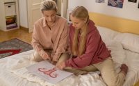 A mother and teenage daughter discuss reproductive health education while sitting on a bed with a schematic diagram.