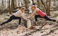 Side view of young female athletes in activewear stretching legs together using fallen tree during fitness workout in spring forest adhering to healthy lifestyle